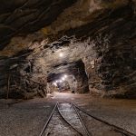 Túnel subterrâneo de uma mina iluminado, com paredes rochosas e trilhos no interior.