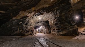 Túnel subterrâneo de uma mina iluminado, com paredes rochosas e trilhos no interior.