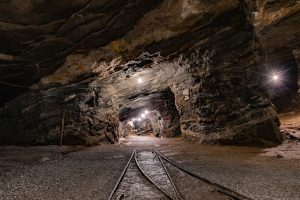 Túnel subterrâneo de uma mina iluminado, com paredes rochosas e trilhos no interior.