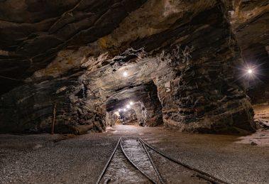 Túnel subterrâneo de uma mina iluminado, com paredes rochosas e trilhos no interior.