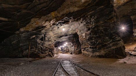Túnel subterrâneo de uma mina iluminado, com paredes rochosas e trilhos no interior.