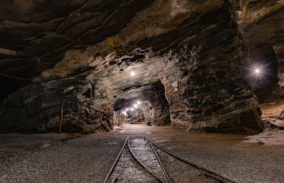 Túnel subterrâneo de uma mina iluminado, com paredes rochosas e trilhos no interior.