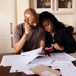 Casal analisa documentos e administra as finanças domésticas enquanto usa um smartphone à mesa.