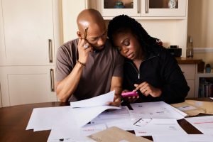 Casal analisa documentos e administra as finanças domésticas enquanto usa um smartphone à mesa.