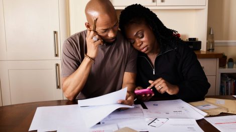 Casal analisa documentos e administra as finanças domésticas enquanto usa um smartphone à mesa.
