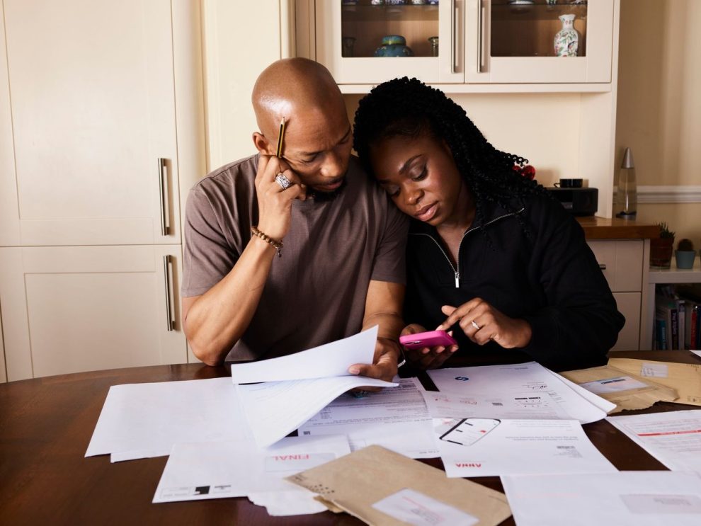 Casal analisa documentos e administra as finanças domésticas enquanto usa um smartphone à mesa.
