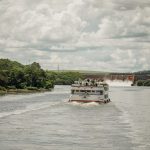 Barco de passageiros navega por um rio cercado por vegetação, próximo a uma usina hidrelétrica.
