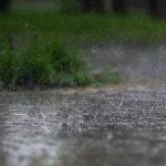 Chuva intensa atingindo o pavimento ao ar livre, com vegetação verde desfocada ao fundo.