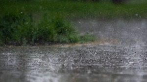 Chuva intensa atingindo o pavimento ao ar livre, com vegetação verde desfocada ao fundo.