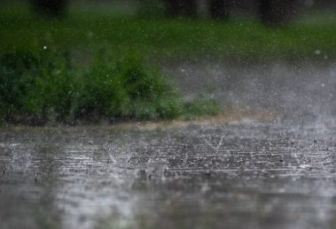 Chuva intensa atingindo o pavimento ao ar livre, com vegetação verde desfocada ao fundo.