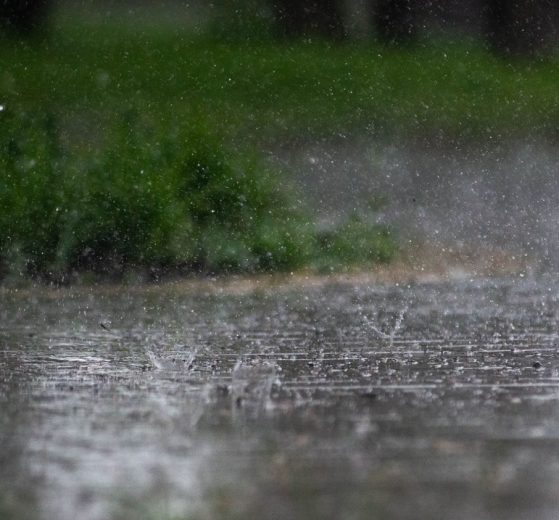 Chuva intensa atingindo o pavimento ao ar livre, com vegetação verde desfocada ao fundo.