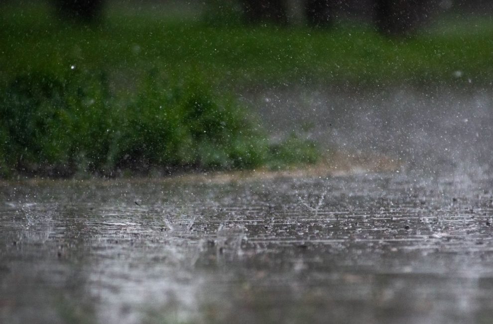 Chuva intensa atingindo o pavimento ao ar livre, com vegetação verde desfocada ao fundo.