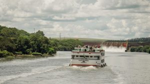 Barco de passageiros navega por um rio cercado por vegetação, próximo a uma usina hidrelétrica.