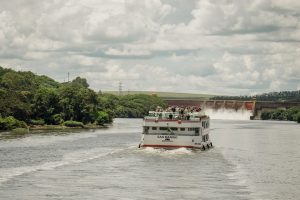 Barco de passageiros navega por um rio cercado por vegetação, próximo a uma usina hidrelétrica.