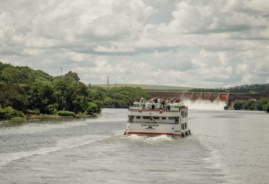 Barco de passageiros navega por um rio cercado por vegetação, próximo a uma usina hidrelétrica.