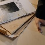Mão apoiada sobre uma mesa próxima a documentos de investigação e recortes de jornal.