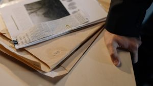 Mão apoiada sobre uma mesa próxima a documentos de investigação e recortes de jornal.