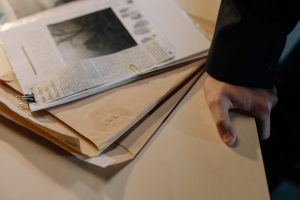 Mão apoiada sobre uma mesa próxima a documentos de investigação e recortes de jornal.