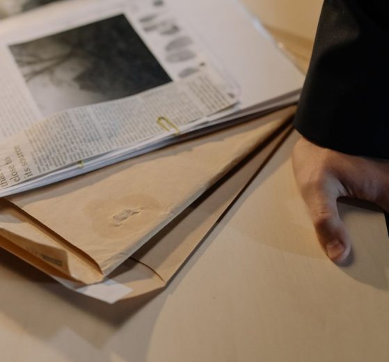 Mão apoiada sobre uma mesa próxima a documentos de investigação e recortes de jornal.