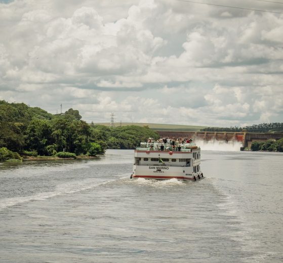 Barco de passageiros navega por um rio cercado por vegetação, próximo a uma usina hidrelétrica.