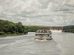 Barco de passageiros navega por um rio cercado por vegetação, próximo a uma usina hidrelétrica.