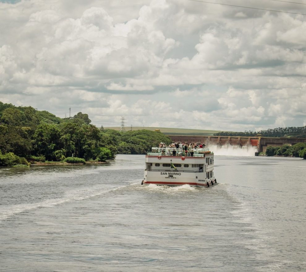 Barco de passageiros navega por um rio cercado por vegetação, próximo a uma usina hidrelétrica.