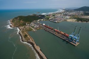 Vista aérea do Porto de Imbituba, com docas, guindastes, navios e a costa brasileira.