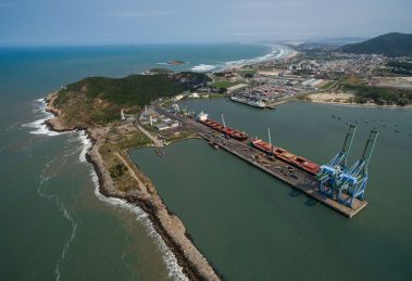 Vista aérea do Porto de Imbituba, com docas, guindastes, navios e a costa brasileira.