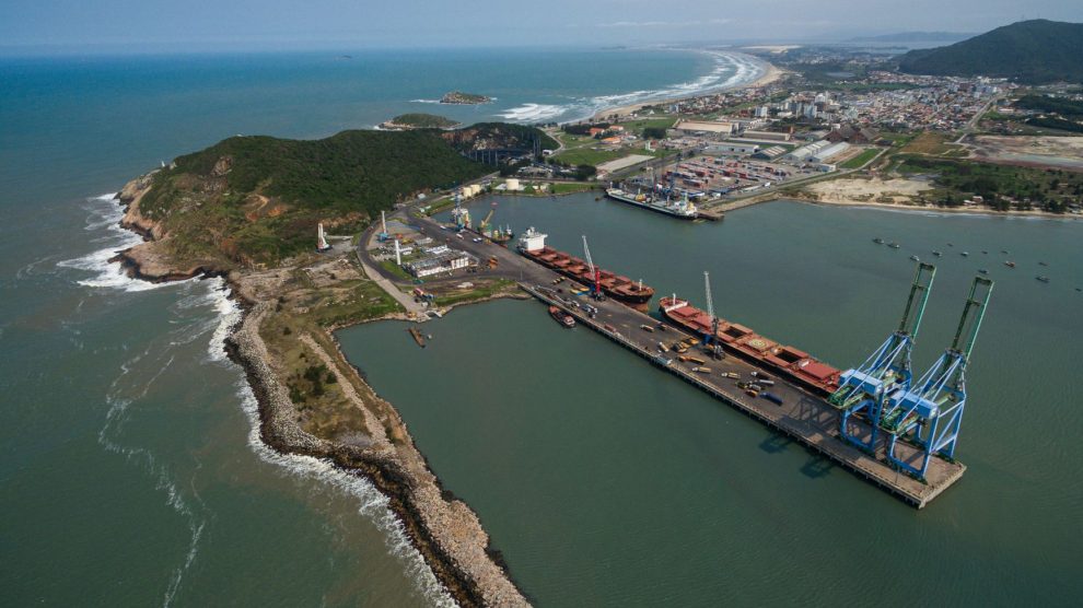 Vista aérea do Porto de Imbituba, com docas, guindastes, navios e a costa brasileira.