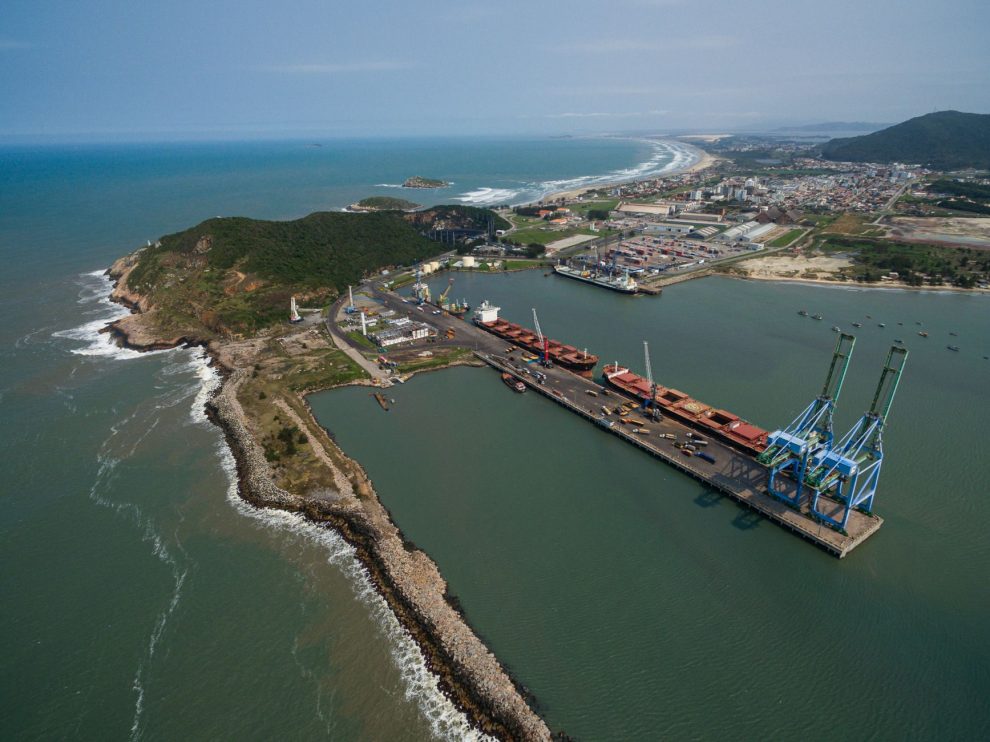 Vista aérea do Porto de Imbituba, com docas, guindastes, navios e a costa brasileira.